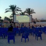 MovieMovil, la empresa de cine al aire libre, crea experiencias mágicas en la playa de El Campello Proyección de cine al aire libre en la playa de El Campello, Alicante, con filas de sillas azules frente a una gran pantalla que muestra información sobre MovieMovil. En el fondo, se observan palmeras, edificios y montañas, mientras el mar completa el paisaje. MovieMovil, la empresa de cine al aire libre