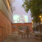 MovieMovil, la empresa de cine al aire libre, ofrece vistas únicas de eventos comunitarios como este partido de fútbol en Ibi Un partido de fútbol proyectado en la calle, en la pared medianera de un edificio en Ibi (Alicante). Se observa un grupo de personas sentadas en mesas y sillas, disfrutando del evento al aire libre. La proyección muestra un partido de alto perfil, con logotipos como Sony y UniCredit visibles en la pantalla. A la izquierda, una pared con graffiti añade un toque urbano, mientras que un árbol a la derecha enmarca la escena. MovieMovil, la empresa de cine al aire libre