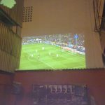 MovieMovil, la empresa de cine al aire libre, lleva la emoción del fútbol a las calles de Ibi para los clientes del bar Proyección de un partido de fútbol en la calle, en la pared medianera de un edificio en Ibi (Alicante), con una gran pantalla mostrando un partido de alto perfil. Se observan logotipos como Sony y UniCredit en la proyección, y el ambiente está diseñado para los clientes de un bar cercano. MovieMovil, la empresa de cine al aire libre