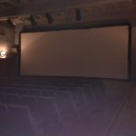 MovieMovil, la empresa de cine al aire libre, crea la atmósfera perfecta para experiencias cinematográficas inolvidables Teatro vacío con asientos enfrentando una gran pantalla blanca, listo para una proyección de cine. La iluminación es suave, y se observan altavoces en las paredes y techo. MovieMovil, la empresa de cine al aire libre