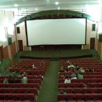 MovieMovil, la empresa de cine al aire libre, transforma la Casa de Cultura de Caudete en un espacio cinematográfico único Proyección de cine en la Casa de Cultura de Caudete, con una gran pantalla blanca situada en el escenario, rodeada de altavoces y filas de asientos rojos dispuestos en niveles. El público está sentado, disfrutando de un ambiente acogedor y cultural. MovieMovil, la empresa de cine al aire libre