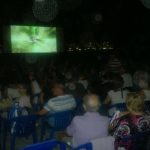 MovieMovil, la empresa de cine al aire libre, crea experiencias mágicas en la playa de El Campello Proyección de cine al aire libre en la playa de El Campello, con una gran pantalla mostrando una película, público sentado en sillas azules y luces de edificios en el fondo. MovieMovil, la empresa de cine al aire libre
