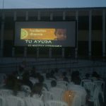 MovieMovil, la empresa de cine al aire libre, apoya causas solidarias con proyecciones como la de la Fundación Dharma en Banyeres de Mariola Proyección de un anuncio de la Fundación Dharma antes de una película en el patio del colegio en Banyeres de Mariola, con una pantalla grande mostrando el texto "Fundación Dharma" y "Tu ayuda es nuestra esperanza", mientras el público observa sentado en sillas blancas. MovieMovil, la empresa de cine al aire libre