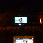 MovieMovil, la empresa de cine al aire libre, combina calidad técnica y emoción en sus proyecciones Proyección de cine al aire libre por la noche, con una gran pantalla mostrando una escena de dos personas abrazándose cerca del mar, público sentado en sillas blancas y un técnico gestionando el audio y video con un portátil en primer plano. MovieMovil, la empresa de cine al aire libre