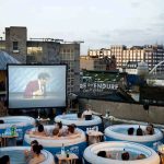 MovieMovil, la empresa de cine al aire libre, crea experiencias exclusivas con proyecciones desde jacuzzis Proyección de cine al aire libre con un enfoque único: los espectadores disfrutan de la película desde jacuzzis inflables, organizados en semicírculo frente a una gran pantalla. Al fondo se observan edificios. MovieMovil, la empresa de cine al aire libre