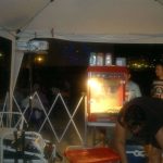 MovieMovil, la empresa de cine al aire libre, cuida cada detalle desde la técnica hasta los sabores Zona de control técnico y palomiteras en un evento de cine al aire libre, con dos máquinas de palomitas iluminadas, un técnico ajustando equipos y público disfrutando del cine en el fondo. MovieMovil, la empresa de cine al aire libre