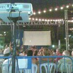 MovieMovil, la empresa de cine al aire libre, honra a los marineros con proyecciones especiales en su día Proyección en un homenaje a los marineros durante el día de su patrona, con un proyector montado en un soporte y una pantalla grande lista para mostrar contenido. El evento tiene lugar al aire libre, bajo luces decorativas y con el público sentado en sillas de plástico. MovieMovil, la empresa de cine al aire libre