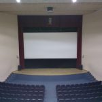 MovieMovil, la empresa de cine al aire libre, transforma la Casa de Cultura de Onil en un espacio cinematográfico único Proyección de cine en la Casa de Cultura de Onil, con una gran pantalla blanca en el escenario, rodeada de altavoces y filas de asientos dispuestas en niveles para el público. MovieMovil, la empresa de cine al aire libre