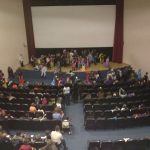MovieMovil, la empresa de cine al aire libre, crea eventos únicos como esta proyección en la Casa de Cultura de Onil Proyección de cine en la Casa de Cultura de Onil, momentos antes de comenzar, con el público disfrazado y un ambiente festivo. Se observa una pantalla grande en el escenario y personas interactuando en sus asientos. MovieMovil, la empresa de cine al aire libre