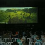 MovieMovil, la empresa de cine al aire libre, adapta sus proyecciones para eventos en interiores con la misma calidad y magia Proyección de cine en una sala cerrada, con público sentado en sillas blancas disfrutando de una película que muestra un paisaje natural con colinas verdes, árboles y un río. MovieMovil, la empresa de cine al aire libre