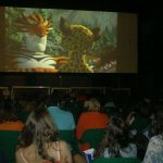 MovieMovil, la empresa de cine al aire libre, transforma la plaza de la iglesia en El Campello en un espacio mágico para disfrutar del cine Proyección de cine al aire libre en la plaza de la iglesia en El Campello, con una gran pantalla mostrando una película animada de un tigre y un leopardo, público sentado en filas de sillas y un ambiente nocturno acogedor. MovieMovil, la empresa de cine al aire libre