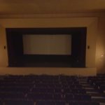 MovieMovil, la empresa de cine al aire libre, transforma teatros emblemáticos en escenarios cinematográficos Teatro vacío listo para una proyección de cine, con filas de asientos de color rojo y un gran escenario con una pantalla blanca. La iluminación es tenue, destacando algunas luces de pared que realzan el ambiente clásico del teatro. MovieMovil, la empresa de cine al aire libre