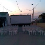 MovieMovil, la empresa de cine al aire libre, transforma las calles de La Gineta en escenarios cinematográficos Proyección de cine al aire libre en una calle de La Gineta, con filas de sillas blancas dispuestas frente a una gran pantalla que muestra una película. El evento tiene lugar en un entorno urbano, con edificios cercanos y luces nocturnas que crean un ambiente acogedor. MovieMovil, la empresa de cine al aire libre