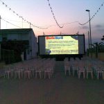 MovieMovil, la empresa de cine al aire libre, transforma las calles de La Gineta en escenarios cinematográficos Proyección de cine al aire libre en una calle de La Gineta, con filas de sillas blancas dispuestas frente a una gran pantalla que muestra información sobre MovieMovil. En el fondo, se observan edificios y luces nocturnas que enmarcan el evento. MovieMovil, la empresa de cine al aire libre