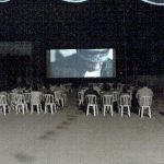 MovieMovil, la empresa de cine al aire libre, transforma las calles de La Gineta en escenarios cinematográficos Proyección de cine al aire libre en una calle de La Gineta, con varias filas de sillas blancas dispuestas frente a una gran pantalla que muestra un primer plano de una persona con sombrero. El evento tiene lugar en un espacio urbano, bajo la iluminación nocturna y rodeado de edificios. MovieMovil, la empresa de cine al aire libre