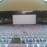 MovieMovil, la empresa de cine al aire libre, transforma el parque "9 de octubre" de Petrer en un espacio cinematográfico comunitario Proyección de cine al aire libre en el parque "9 de octubre" de Petrer, con una gran pantalla blanca instalada en un escenario bajo una estructura cubierta. Filas de sillas blancas están dispuestas para el público, rodeadas de un entorno verde con árboles y césped. MovieMovil, la empresa de cine al aire libre