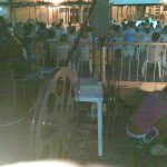 MovieMovil, la empresa de cine al aire libre, revive la magia del cine en 35mm en Beniel Proyección de cine en 35mm en Beniel, con un proyector clásico y una gran pantalla mostrando una película. Se observa un carrete de película junto al proyector, mientras el público disfruta sentado en un espacio al aire libre. MovieMovil, la empresa de cine al aire libre