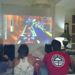 MovieMovil, la empresa de cine al aire libre, transforma espacios privados en salas de videojuegos únicas Proyección de videojuegos en una casa privada, con un grupo de personas disfrutando de un juego de carreras futurista proyectado en una pantalla grande. La sala está equipada con un sofá, mesa, lámparas y altavoces, creando un ambiente cómodo y social. MovieMovil, la empresa de cine al aire libre
