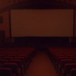 MovieMovil, la empresa de cine al aire libre, transforma la Casa de Cultura de Caudete en un espacio cinematográfico único Interior del cine en la Casa de Cultura de Caudete, con filas de asientos rojos dispuestos en niveles, todos orientados hacia una gran pantalla blanca situada en el escenario. El espacio está iluminado de manera tenue, creando un ambiente acogedor y típico de una sala de cine. MovieMovil, la empresa de cine al aire libre