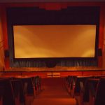 MovieMovil, la empresa de cine al aire libre, transforma el teatro de Xixona en un espacio cinematográfico único Interior del teatro de Xixona (Alicante), con filas de asientos rojos dispuestos en niveles, todos orientados hacia una gran pantalla blanca situada en el escenario. Las paredes del teatro son de color rojo, con altavoces montados en ellas, y el suelo es de madera, creando un ambiente cálido y acogedor. MovieMovil, la empresa de cine al aire libre