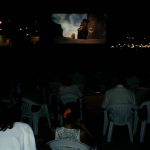 MovieMovil, la empresa de cine al aire libre, transforma la playa en un escenario mágico para disfrutar del cine Proyección de cine al aire libre en una playa, con una gran pantalla mostrando una película animada, posiblemente de la saga "Ice Age", que incluye un mamut y un tigre dientes de sable. El público está sentado en sillas de plástico dispuestas en filas, disfrutando del evento bajo el cielo nocturno. MovieMovil, la empresa de cine al aire libre