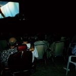 MovieMovil, la empresa de cine al aire libre, transforma la playa en un escenario mágico para disfrutar del cine Proyección de cine al aire libre en una playa, con una gran pantalla blanca mostrando una película animada que incluye un tigre dientes de sable. El público está sentado en filas de sillas, disfrutando del evento en un ambiente relajado bajo el cielo nocturno y con el sonido del mar como fondo. MovieMovil, la empresa de cine al aire libre