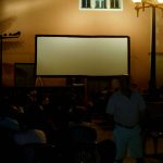 MovieMovil, la empresa de cine al aire libre, transforma la plaza de la iglesia de El Campello en un espacio mágico para disfrutar del cine Proyección de cine al aire libre en la plaza de la iglesia de El Campello, con una gran pantalla montada en la pared y un público sentado en sillas, esperando el inicio de la película. La iluminación nocturna y un farol cercano añaden un toque acogedor al evento. MovieMovil, la empresa de cine al aire libre