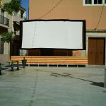 MovieMovil, la empresa de cine al aire libre, transforma la plaza de la iglesia de El Campello en un espacio mágico para disfrutar del cine Proyección de cine al aire libre en la plaza de la iglesia en El Campello, Alicante, con una gran pantalla blanca bordeada en negro, elevada sobre una plataforma decorada con franjas amarillas y naranjas. El entorno incluye edificios cercanos, uno con balcón y banderas, además de luces decorativas suspendidas sobre la pantalla y una farola que proyecta sombras en el suelo. MovieMovil, la empresa de cine al aire libre