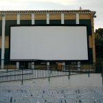 MovieMovil, la empresa de cine al aire libre, transforma Banyeres de Mariola en un espacio mágico para disfrutar del cine Proyección de cine al aire libre en Banyeres de Mariola, con una gran pantalla blanca montada en la pared de un edificio de ladrillo con columnas blancas. Filas de sillas blancas están dispuestas para el público, y se observa un entorno verde con árboles en el fondo. MovieMovil, la empresa de cine al aire libre