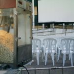 MovieMovil, la empresa de cine al aire libre, transforma Banyeres de Mariola en un escenario perfecto para disfrutar del cine royección de cine al aire libre en Banyeres de Mariola, con una máquina de palomitas en primer plano llena de palomitas listas para servir. Al fondo, se observan filas de sillas blancas dispuestas frente a una gran pantalla blanca, creando un ambiente acogedor y comunitario. MovieMovil, la empresa de cine al aire libre