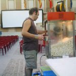 MovieMovil, la empresa de cine al aire libre, transforma la plaza del Ayuntamiento de Elda en un espacio mágico para disfrutar del cine Proyección de cine al aire libre en la plaza del Ayuntamiento de Elda, con un hombre operando una máquina de palomitas en primer plano. Al fondo, se observan filas de sillas rojas dispuestas frente a una gran pantalla blanca, creando un ambiente comunitario y acogedor. MovieMovil, la empresa de cine al aire libre