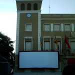 MovieMovil, la empresa de cine al aire libre, transforma la plaza del Ayuntamiento de Elda en un espacio mágico para disfrutar del cine Proyección de cine al aire libre en la plaza del Ayuntamiento de Elda, con una gran pantalla blanca instalada frente al edificio del Ayuntamiento, que cuenta con una torre con reloj y varias banderas colgando. Filas de sillas están dispuestas para el público, creando un ambiente comunitario y acogedor. MovieMovil, la empresa de cine al aire libre