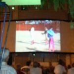 MovieMovil, la empresa de cine al aire libre, rinde homenaje a un torero en un evento especial en Petrer Proyección de cine al aire libre en una plaza de Petrer, homenajeando a un torero frente a la puerta de su casa, con una pantalla grande mostrando imágenes taurinas y público sentado observando. MovieMovil, la empresa de cine al aire libre