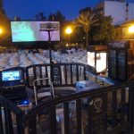 MovieMovil, la empresa de cine al aire libre, combina proyección, palomitas y emoción bajo las estrellas Montaje de cine al aire libre con pantalla grande proyectando una imagen colorida, sillas blancas para el público, una zona técnica con proyector y equipos, y una máquina de palomitas y Coca-Cola. MovieMovil, la empresa de cine al aire libre