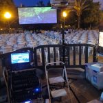 MovieMovil, la empresa de cine al aire libre, presenta su impecable montaje técnico para proyecciones únicas Texto alternativo: Proyección de cine al aire libre con una pantalla grande, sillas de plástico blanco dispuestas para el público, un proyector en un soporte y equipo técnico como un portátil y mezclador de sonido. MovieMovil, la empresa de cine al aire libre