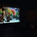 MovieMovil ofrece noches inolvidables de cine al aire libre, con proyecciones animadas para disfrutar bajo las estrellas. Proyección nocturna de cine al aire libre con pantalla que muestra una escena animada en un parque, organizada por MovieMovil.