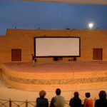 MovieMovil, la empresa de cine al aire libre, transforma espacios urbanos en escenarios cinematográficos únicos Proyección de cine al aire libre en Ibi, con una gran pantalla blanca instalada en la pared de un edificio de ladrillo. En el escenario elevado, se observa una pared de piedra con graffiti que añade un toque urbano al entorno. Varias personas están sentadas en el área del público, disfrutando del evento al aire libre. MovieMovil, la empresa de cine al aire libre