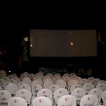 MovieMovil, la empresa de cine al aire libre, ofrece noches de cine inolvidables bajo las estrellas Proyección de cine al aire libre con un gran pantalla situada frente a filas de sillas blancas, cada una con logos o texto en la parte posterior, indicando patrocinadores o branding del evento. El lugar está decorado con carteles o pancartas que flanquean la pantalla, y la iluminación nocturna crea un ambiente acogedor y comunitario. MovieMovil, la empresa de cine al aire libre