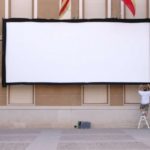 MovieMovil, la empresa de cine al aire libre, prepara la plaza del Ayuntamiento de Elda para una noche mágica de cine Montaje de una pantalla de cine al aire libre en la plaza del Ayuntamiento de Elda, con una persona en una escalera ajustando la pantalla blanca instalada en la fachada del edificio. El entorno incluye banderas y detalles arquitectónicos que reflejan el carácter público del espacio. MovieMovil, la empresa de cine al aire libre