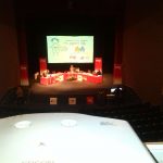 MovieMovil, la empresa de cine al aire libre, colabora en eventos solidarios como la campaña "Un juguete, una ilusión" Proyección durante un programa de Radio Nacional de España en la campaña "Un juguete, una ilusión" en la Casa de Cultura de Onil, con una pantalla grande mostrando un dibujo de un niño con un juguete, mesas con manteles rojos y equipo técnico en el escenario. MovieMovil, la empresa de cine al aire libre