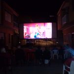 MovieMovil, la empresa de cine al aire libre, lleva la magia del cine a espacios únicos y acogedores Proyección de cine al aire libre en una calle estrecha, con una pantalla grande mostrando una película animada, público sentado en sillas observando, y edificios alrededor creando una atmósfera íntima. MovieMovil, la empresa de cine al aire libre