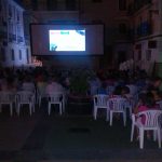 MovieMovil, la empresa de cine al aire libre, reúne a la comunidad con proyecciones al aire libre Proyección al aire libre organizada por MovieMovil, mostrando un anuncio de la empresa en una pantalla grande, con público sentado en sillas blancas, en un espacio abierto rodeado de edificios. MovieMovil, la empresa de cine al aire libre