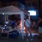 MovieMovil, la empresa de cine al aire libre, combina tecnología y sabor con palomitas y refrescos Zona de control técnico y venta de palomitas y refrescos en un evento de cine al aire libre, con equipo técnico, máquina de palomitas y contenedores de refrescos bajo una carpa iluminada. MovieMovil, la empresa de cine al aire libre