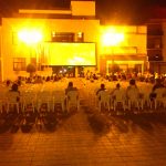MovieMovil, la empresa de cine al aire libre, transforma la plaza del Ayuntamiento de Catral en un espacio cultural único Proyección de cine al aire libre en la plaza del Ayuntamiento de Catral, con una gran pantalla mostrando una película, sillas blancas para el público y edificios emblemáticos alrededor. MovieMovil, la empresa de cine al aire libre