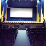 MovieMovil, la empresa de cine al aire libre, organiza proyecciones impecables en espacios emblemáticos como el Auditorio ADOC Preparativos finales para una proyección de cine en el Auditorio ADOC de Elda, con una pantalla grande, cortinas azules, filas de asientos y equipo técnico en el escenario. MovieMovil, la empresa de cine al aire libre