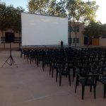 MovieMovil la empresa del cine al aire libre transforma un colegio en Aspe en un espacio mágico para disfrutar del cine bajo las estrellas. Preparativos finales para una proyección de cine al aire libre en un colegio en Aspe, organizada por MovieMovil la empresa del cine al aire libre, con pantalla de 9,5m x 5m y sillas negras dispuestas para los asistentes.