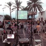 MovieMovil la empresa del cine al aire libre transforma Altea en un escenario mágico para disfrutar del cine bajo las estrellas. Proyección de cine al aire libre en Altea, organizada por MovieMovil la empresa del cine al aire libre, con pantalla de 4m x 3m, sillas blancas y un entorno pintoresco con palmeras y edificios tradicionales.