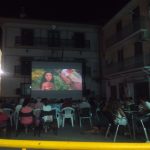 MovieMovil la empresa del cine al aire libre transforma espacios rodeados de historia en escenarios mágicos para disfrutar del cine. Proyección de cine al aire libre en un patio rodeado de edificios con balcones y un reloj visible, organizada por MovieMovil la empresa del cine al aire libre, con pantalla gigante y ambiente comunitario nocturno.