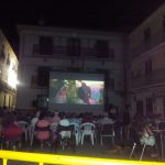 MovieMovil la empresa del cine al aire libre transforma espacios rodeados de historia en escenarios mágicos para disfrutar del cine. Proyección de cine al aire libre en un patio rodeado de edificios con balcones y un reloj visible, organizada por MovieMovil la empresa del cine al aire libre, con pantalla gigante y un ambiente comunitario nocturno.