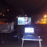 MovieMovil la empresa del cine al aire libre transforma la playa de El Campello en un escenario mágico para disfrutar del cine bajo las estrellas. Proyección de cine al aire libre en la playa de El Campello, organizada por MovieMovil la empresa del cine al aire libre. El evento incluye una pantalla gigante, un puesto de palomitas y un ambiente nocturno junto al mar, con edificios iluminados al fondo.